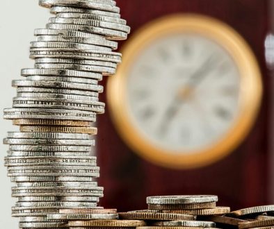 A close-up image of stacked coins with a blurred clock, symbolizing time and money relationship.