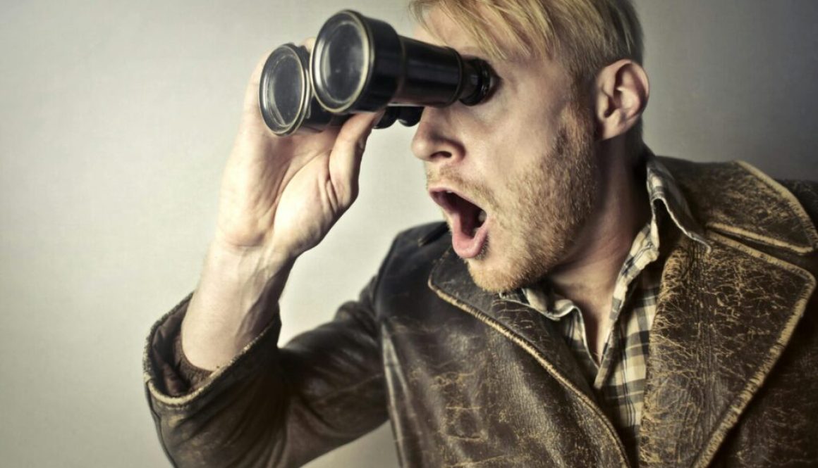 A surprised man in a leather jacket peers through binoculars with a shocked expression.