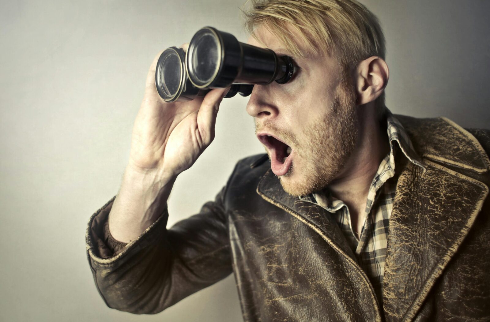A surprised man in a leather jacket peers through binoculars with a shocked expression.